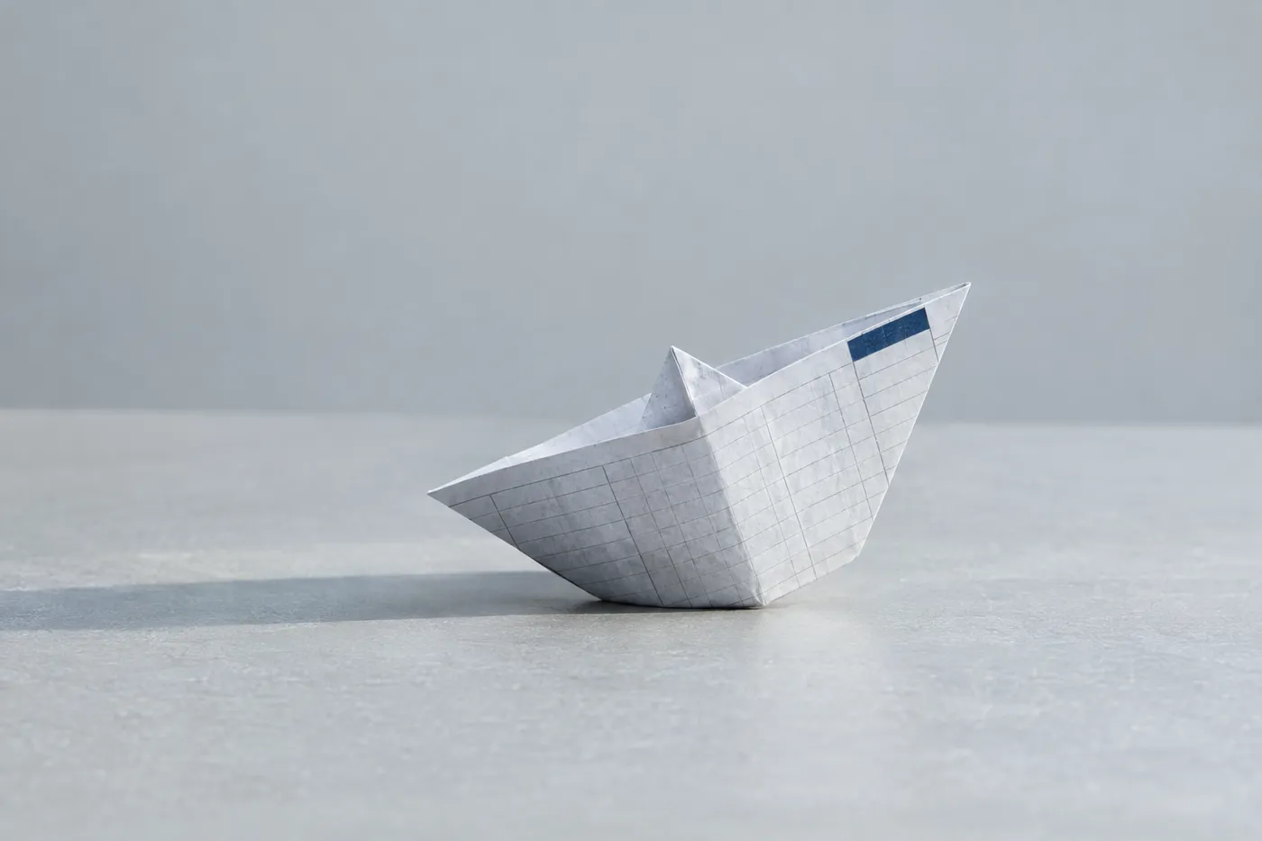 A paper boat folded from ledger paper tilts precariously on a grey surface, representing a sinking accountant cover letter.