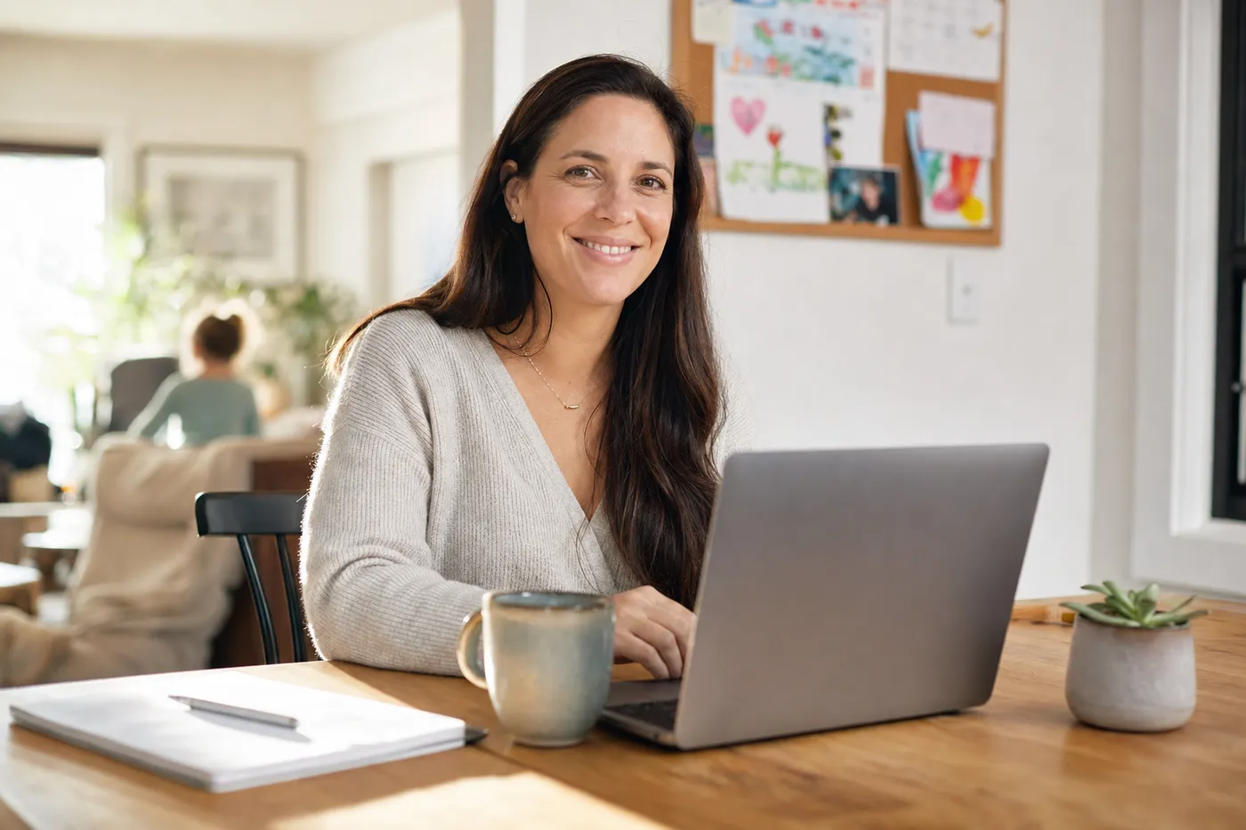 A woman smiling confidently at her sunlit home workspace, preparing her return to the professional workforce.