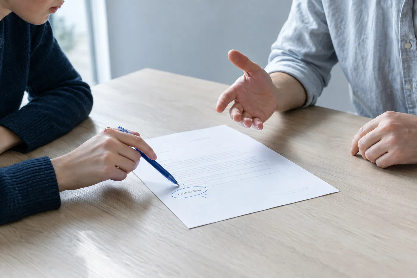 A hiring manager points a blue pen to a circled link on a cover letter during a candid interview with a video editor.