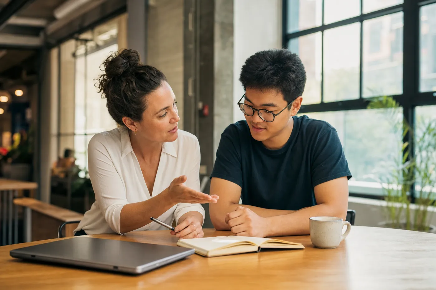 A mentor and a young professional discuss resume formatting options together at a sunlit wooden table.