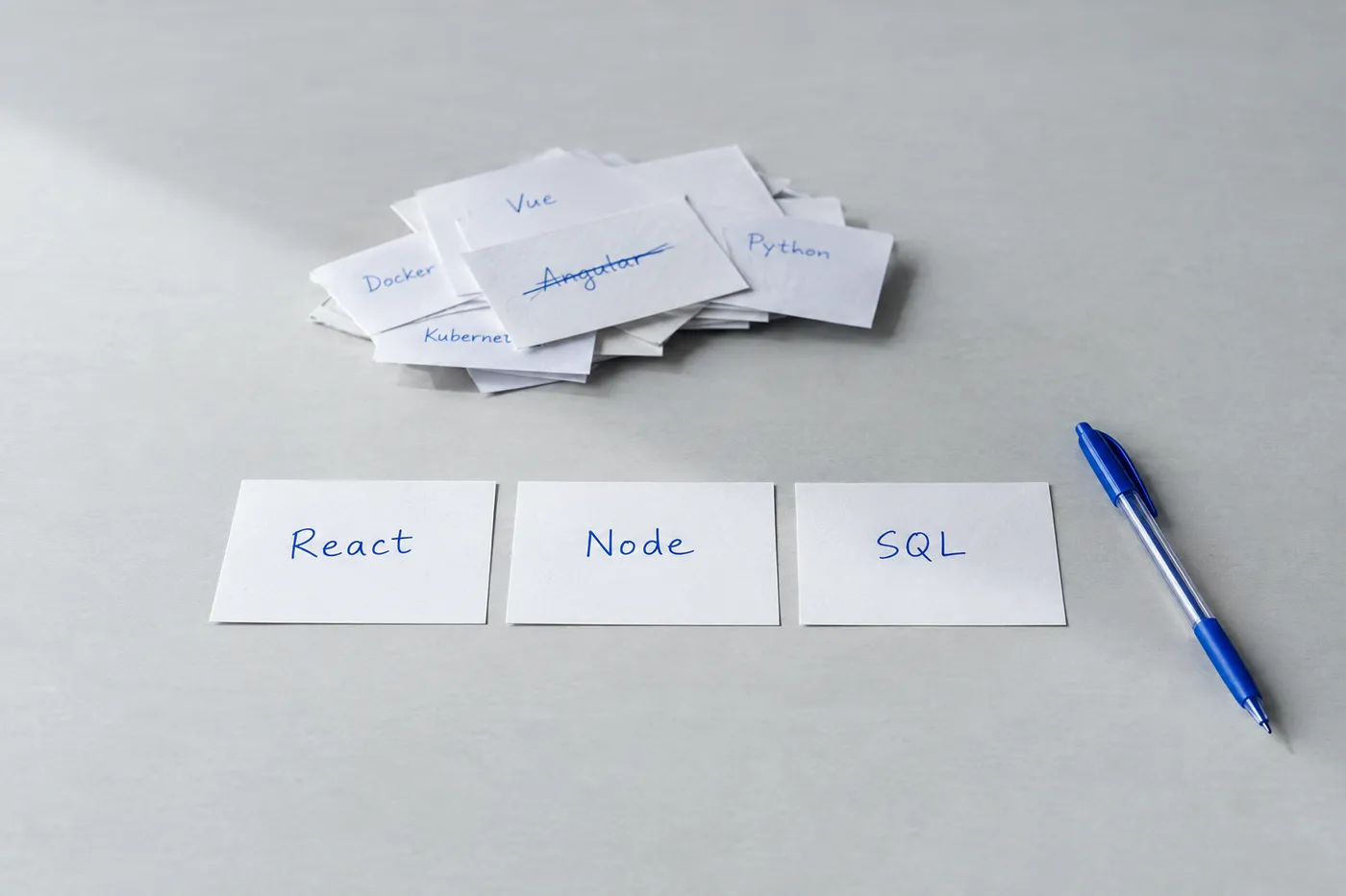 Three neat index cards labeled React, Node, and SQL on a desk, with other discarded technology cards pushed aside.
