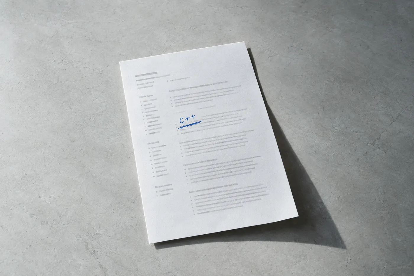 A printed resume on a grey surface with the word C++ handwritten in blue ink over a crossed-out line.