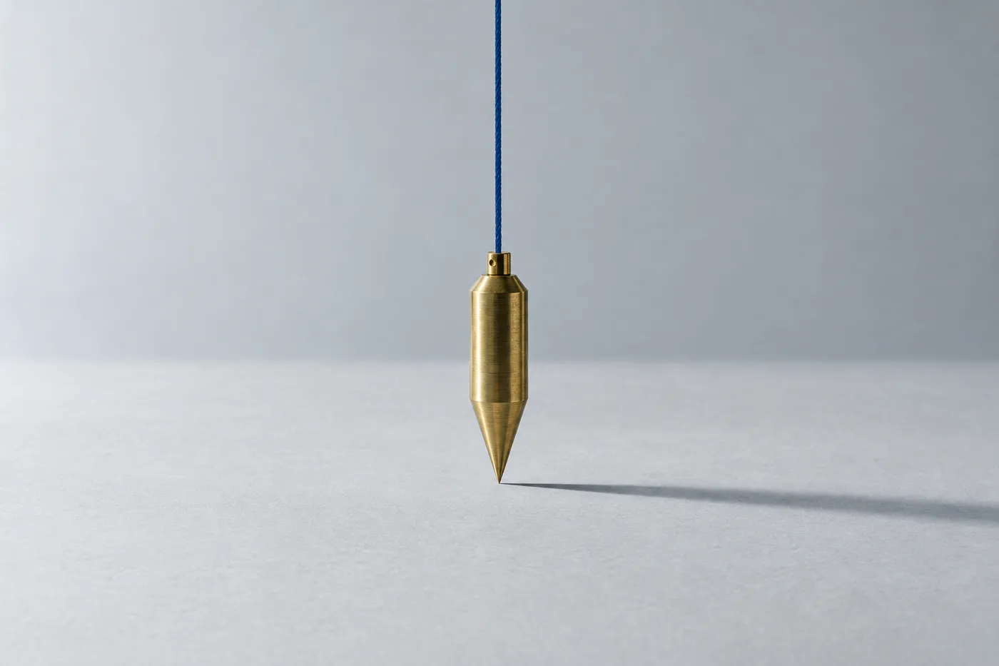 A brass plumb bob hangs suspended over a grey surface, casting a sharp shadow that illustrates perfect vertical alignment.